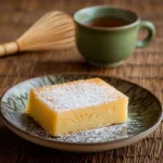 Freshly baked butter mochi slice on a traditional Japanese plate with powdered sugar, green tea cup, and bamboo whisk.